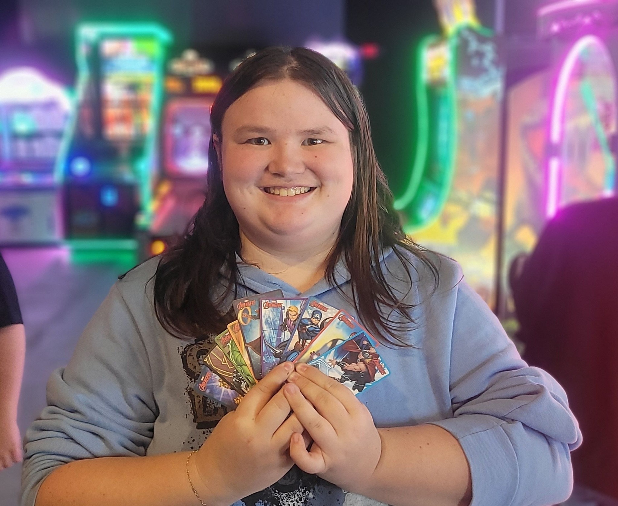 A smiling young person holds a fan of Marvel Avengers-themed trading cards in front of an arcade game.