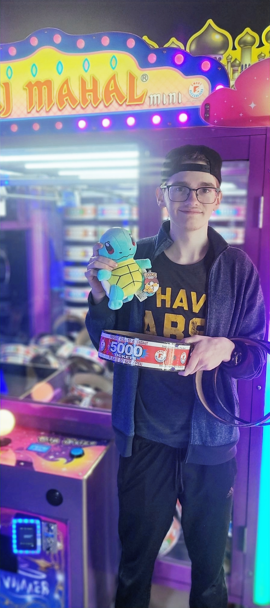 A young man wearing glasses and a cap stands in front of a colorful arcade claw machine, smiling while holding a Squirtle plush toy.