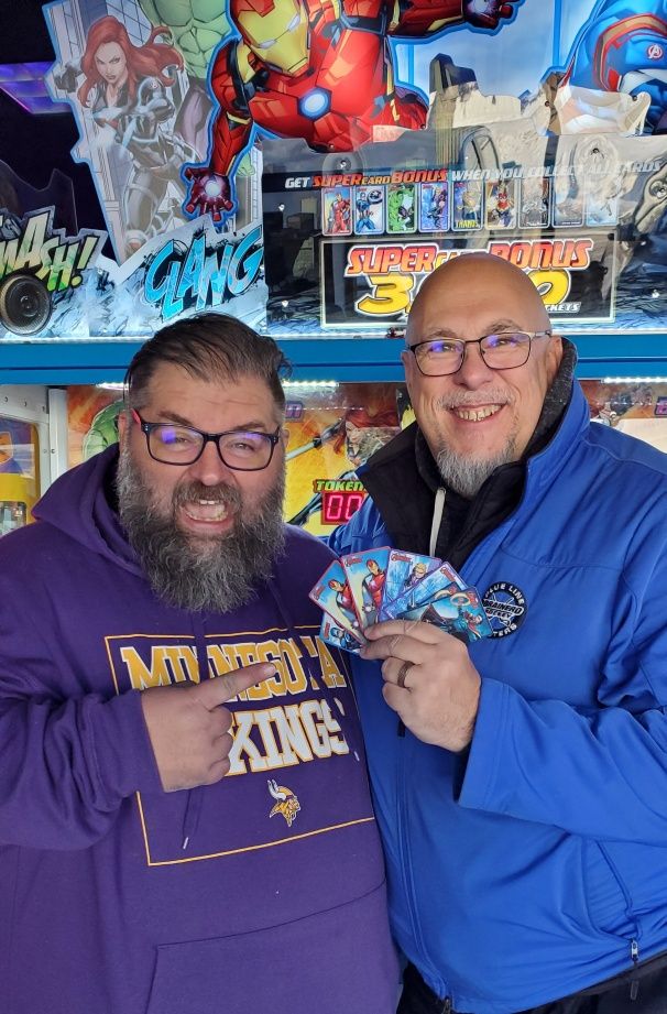 Two men pose in front of an Avengers arcade pusher game.