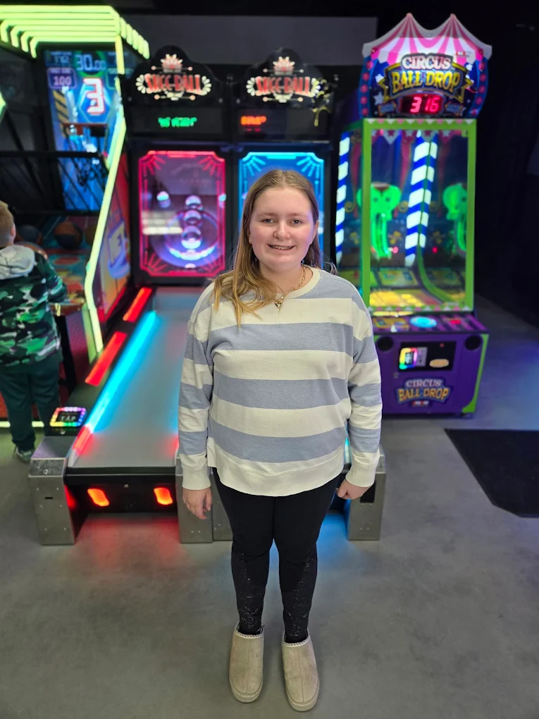 A young girl stands smiling in front of Skee-Ball and Circus Ball Drop arcade games.