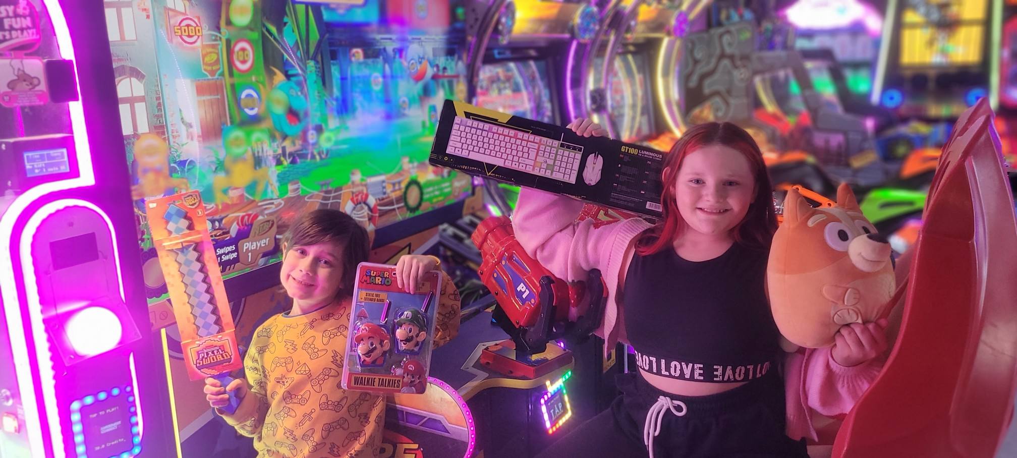 Two children smile with arcade prizes in the brightly lit arcade.