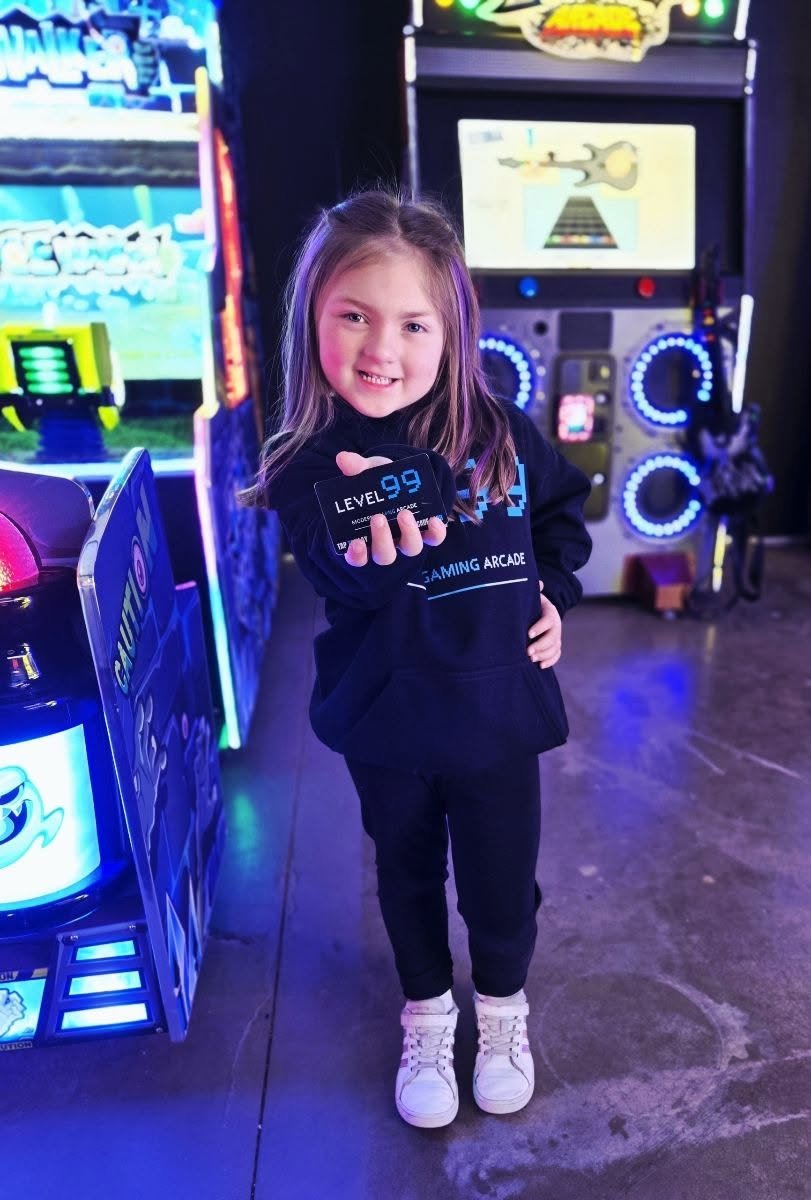 A smiling girl holds up her Level 99 game card in front of glowing arcade games
